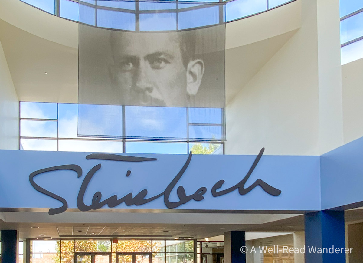The word Steinbeck in handwriting underneth a hanging portrait of Steinbeck in the entryway of a building with natural light