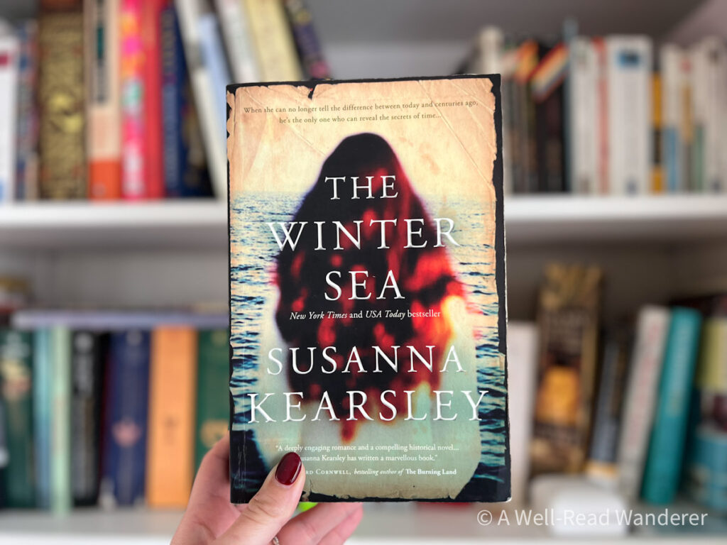 the winter sea by susanna kearsley, book cover with back of redhead woman's head. book held in front of bookshelf
