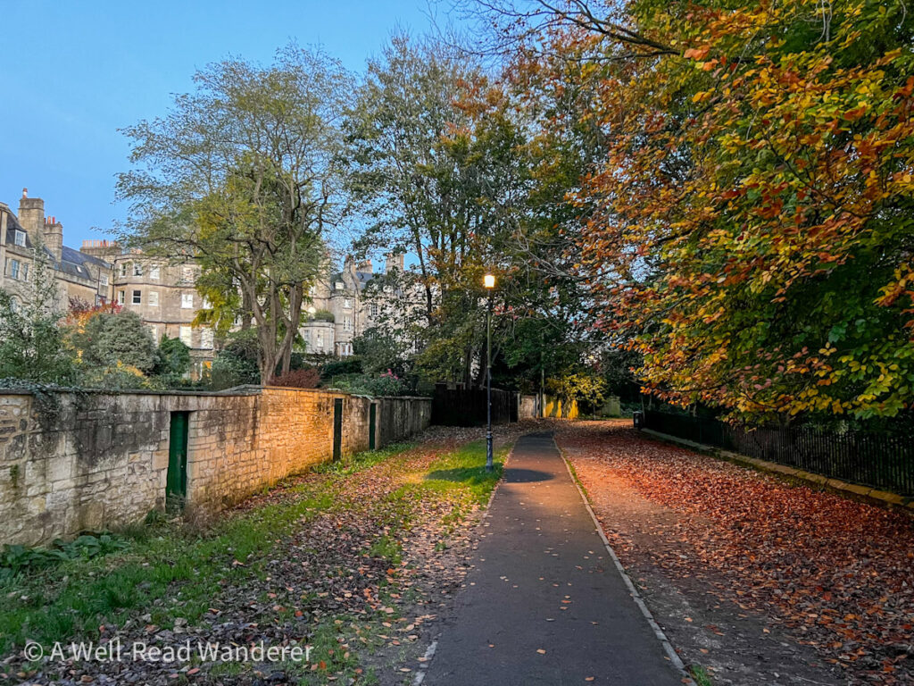 at dusk a lamp illuminated gravel walk in bath uk with trees and georgian buildings in the distance, a scene from a jane austen novel