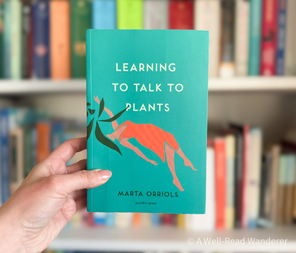 learning to talk to plants catalan novel held by manicured hand in front of white bookshelf